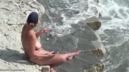 Курящая голая блондинка с телефоном сидит на берегу моря